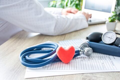 A doctor works on reports with his medical tools next to him on his desk, in front of a red healthy heart toy